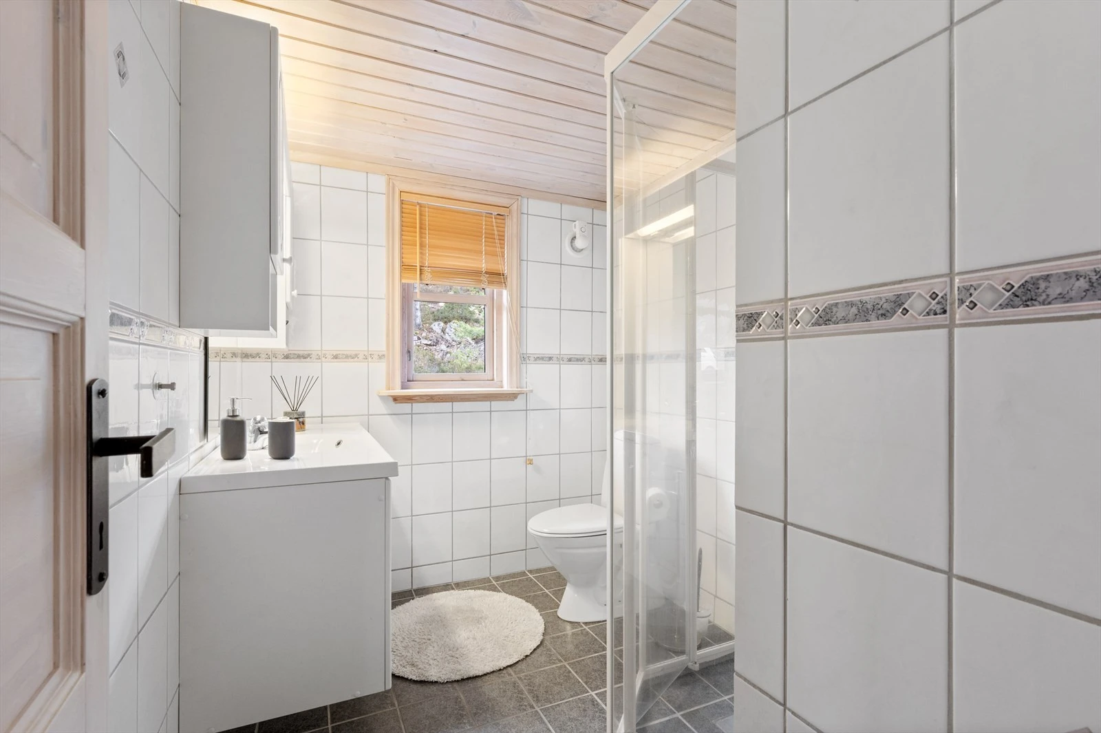 Pent flislagt bad med dusj, helstøpt servant i baderomsinnredning og toalett. Galleribilde