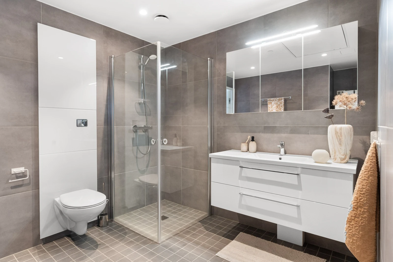 Lekkert og større baderom som inneholder dusj med innfellbare dører i herdet glass, servant, veggfestet klosset, samt benkeskap og veggskap. Galleribilde