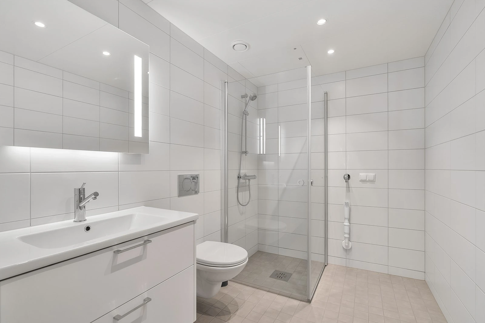 Moderne bad med dusjhjørne, vegghengt toalett med soft-close sete, og stilrene fliser i grå keramikk på gulv og lyse fliser på veggene.