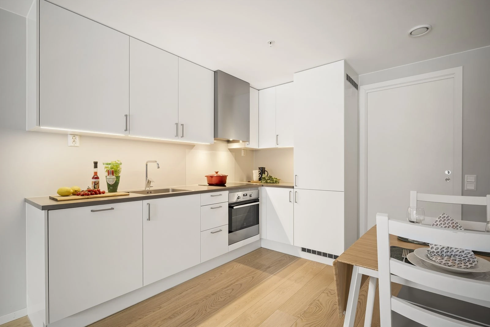 Vegg over kjøkkenbenk med glassplate ved platetopp og malte flater, samt. frittstående hvitevarer.