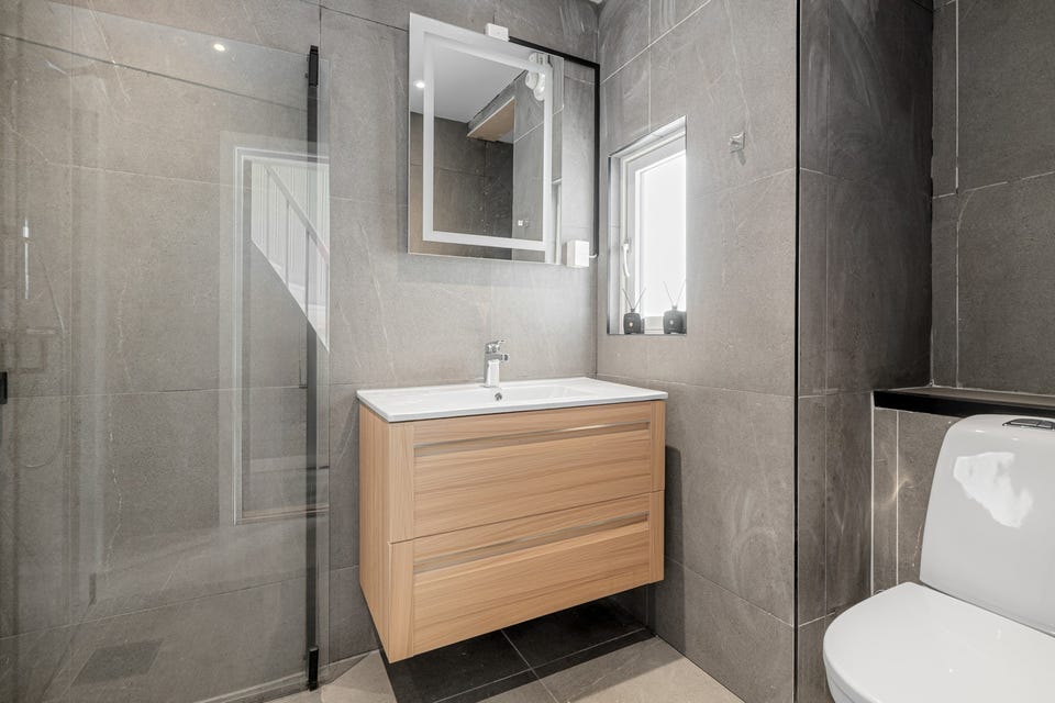 Badet ligger lett tilgjengelig fra entréen og inneholder dusj med innfellbare glassdører, servant med underskap, toalett og speil med lys.