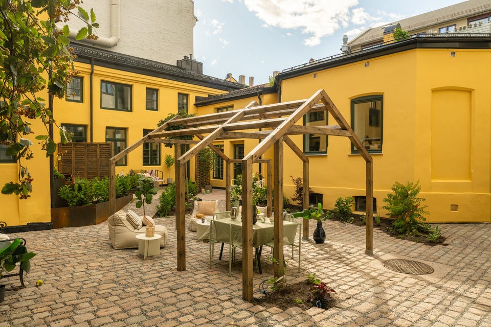 Felles pergola med plass til sittegruppe, omgitt av brostein og gul murfasade. Møblene på bildet er styling.