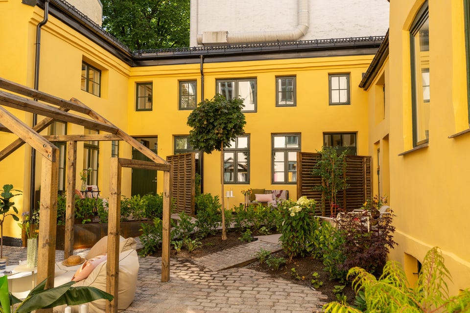Felles gårdsrom sett mellom pergolaen og den gule bakgårdsfasaden. Private terrasser langs bygget tilhører andre leiligheter.