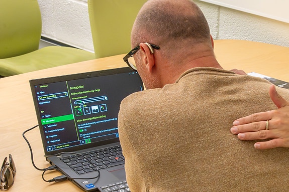 En person med briller og høreapparat foran en PC med forstørrelsesprogram