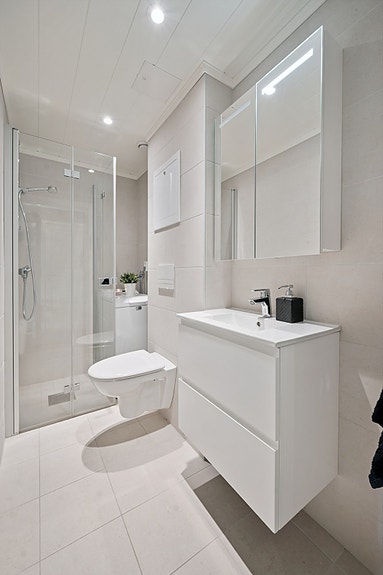 Rommet har innredning med nedfelt servant med skuffer, speilskap, veggmontert toalett, dusjvegger/hjørne og opplegg for vaskemaskin.