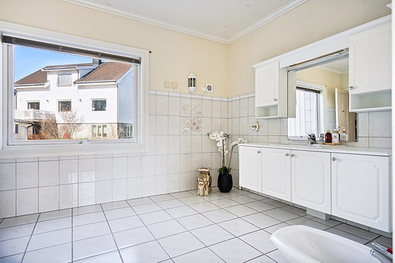 Badet i 1. etasje har fliser og malte plater på vegger og fliser på gulv. Det er innredning med nedfelt servant, gulvmontert toalett, dusjkabinett, bide og opplegg for vaskemaskin.