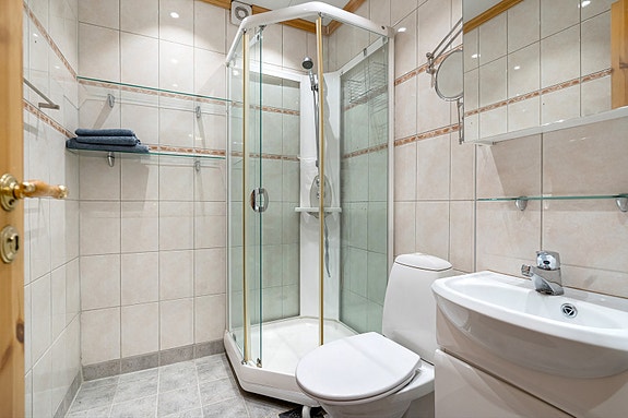 Badet har fliser på både gulv og vegg, dusjkabinett, toalett og servant med underskap