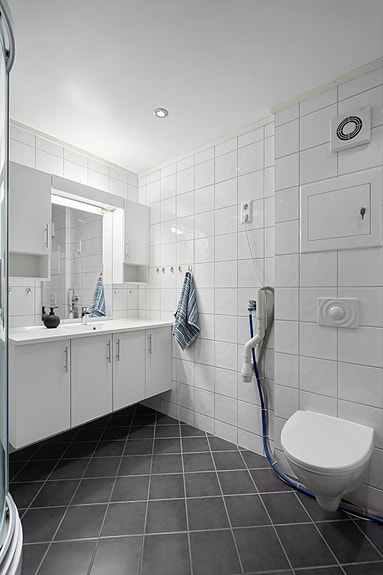 Stort flislagt bad i underetasje med vegghengt toalett, servant med skap, speil, overskap og dusjkabinett