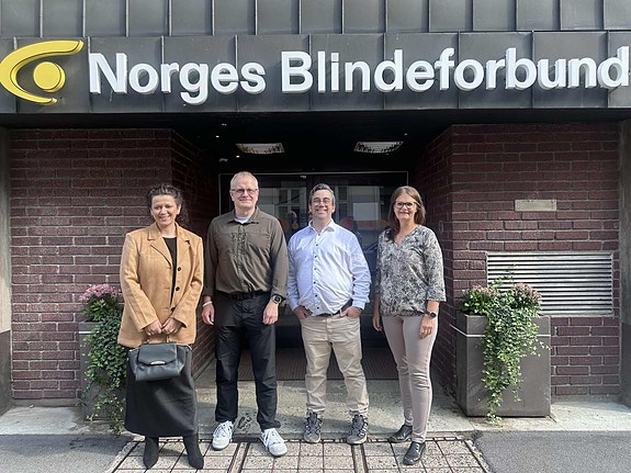 Fire personer står samlet og smiler foran en mursteinsfasade. Over inngangsdøren henger skiltet «Norges Blindeforbund». Det står blomsterkasser på hver side av døren.