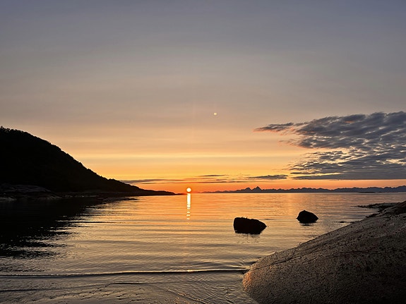 Midnattsol ved sjøen, med solen lavt over horisonten som speiler seg i rolig vann. Steiner i forgrunnen og fjell i bakgrunnen rammer inn landskapet. Bilde tatt av en ansatt.