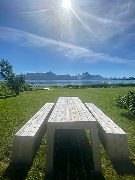 Uteplass hos KUN med benk og bord på en grønn gressplen ved sjøen. Sollys, rolig vann og fjell i bakgrunnen skaper en fredelig uteplass. Bilde tatt av en ansatt.