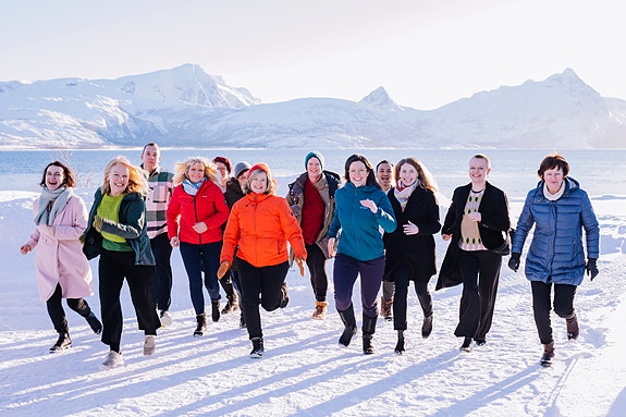 Ansatte på KUN løper smilende mot kamera i et snødekt landskap med fjell og vann i bakgrunnen. Foto: Karoline O.A. Pettersen (2022).
