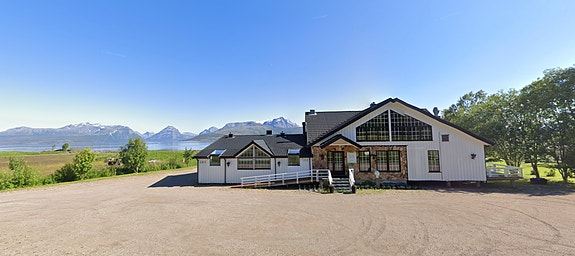 KUN-bygget i Steigen, en hvitmalt bygning med universelt utformet inngangsparti og store vinduer, ligger i et åpent landskap. Utsikt mot hav og fjell i bakgrunnen.